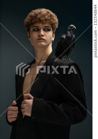 young man with curly hair and a bare torso stands confidently in a black jacket a raven resting on his shoulder. The indoor lighting creates a moody atmosphere around him. young man with curly hair and a bare torso stands confidently in a black jacket a raven resting on his shoulder. The indoor lighting creates a moody atmosphere around him. 132548644