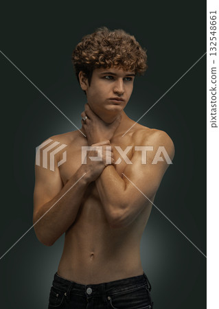 Curly haired young man poses with a thoughtful expression gently holding his neck against a dark background. The soft lighting highlights his torso and features creating a striking visual. Curly haired young man poses with a thoughtful expression gently holding his neck against a dark background. The soft lighting highlights his torso and features creating a striking visual. 132548661
