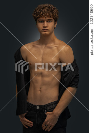 young man stands in a studio showcasing his curly hair and toned physique. He wears a black shirt casually draped over one shoulder while looking directly at the camera. young man stands in a studio showcasing his curly hair and toned physique. He wears a black shirt casually draped over one shoulder while looking directly at the camera. 132548690