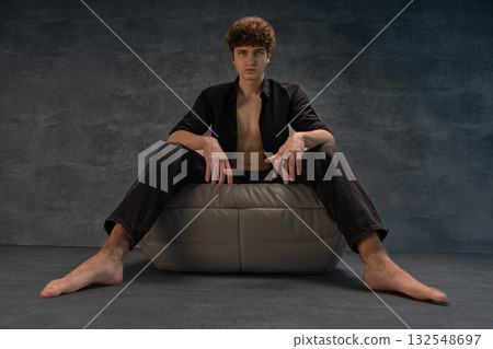 young man with curly hair sits comfortably on a soft cushion in a studio. He wears a black shirt and jeans striking a relaxed yet confident pose with bare feet. young man with curly hair sits comfortably on a soft cushion in a studio. He wears a black shirt and jeans striking a relaxed yet confident pose with bare feet. 132548697