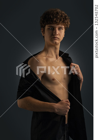 young man with curly hair stands confidently in a studio with a dark background. He wears a black shirt partially unbuttoned revealing his bare chest. The lighting highlights his features. 132548702
