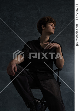 young man with curly hair sits confidently in a black shirt leaning slightly on a chair. The studio backdrop adds depth emphasizing his relaxed pose and contemplative mood. 132548711