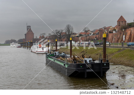 Harbor scene in Tangermuende with boats and barge moored along Elbe River under cloudy winter sky. Historic brick walls and medieval towers create a quiet atmosphere of timeless riverside town. 132548712