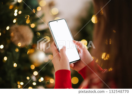 A close-up of a person's hands in a red sweater holding a vertical smartphone with a bright white blank screen against a background of sparkling Christmas bokeh. A close-up of a person's hands in a red sweater holding a vertical smartphone with a bright white blank screen against a background of sparkling Christmas bokeh. 132548770