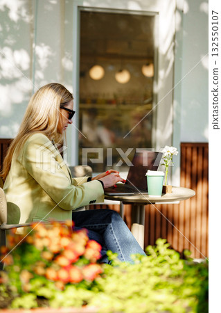 Woman working on laptop at outdoor cafe surrounded by plants and flowers in warm sunlight Woman working on laptop at outdoor cafe surrounded by plants and flowers in warm sunlight 132550107