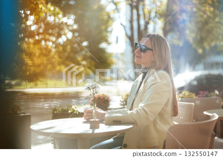 Young woman enjoying sunny day outside cafe with drink in hand and flowers on table Young woman enjoying sunny day outside cafe with drink in hand and flowers on table 132550147