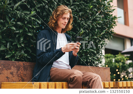 Young man checks his phone while sitting on a bench in an urban setting during daytime Young man checks his phone while sitting on a bench in an urban setting during daytime 132550150