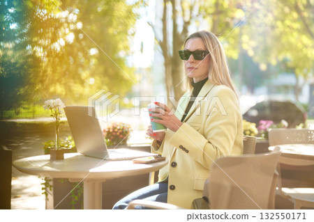 Young woman working on laptop outside while enjoying coffee at a sunny cafe 132550171