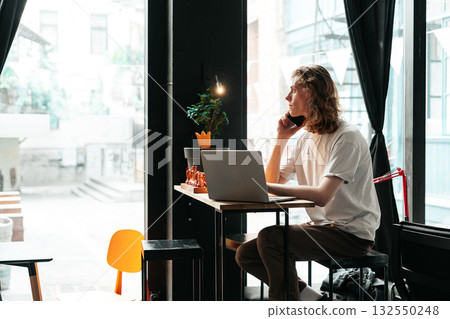 Man sitting at a table in a cafe, talking on his phone while working on a laptop 132550248