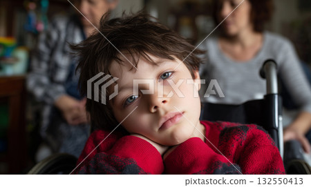 Young boy with disability looking at camera, sitting in a wheelchair, family members caring in background Young boy with disability looking at camera, sitting in a wheelchair, family members caring in background 132550413