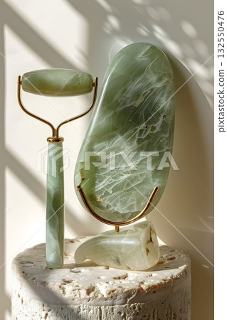 Minimal beauty setup featuring a polished jade gua-sha board, facial roller on a gold stand, oval massage stone and jade wand arranged on a textured stone tray. Clean light and marble veins emphasize  132550476