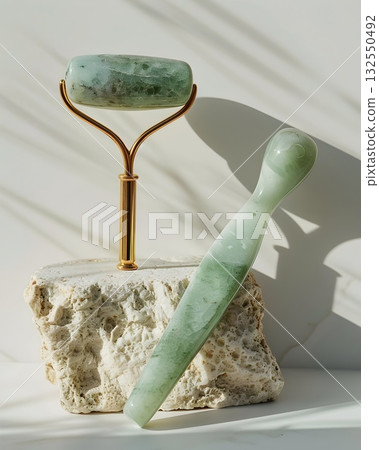 Minimal beauty setup featuring a polished jade gua-sha board, facial roller on a gold stand, oval massage stone and jade wand arranged on a textured stone tray. Clean light and marble veins emphasize  132550492