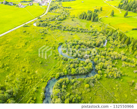 A tranquil river winds gracefully through a verdant valley, surrounded by lush greenery and dotted with trees. The stunning alpine landscape reveals nature's beauty from above. 132550966