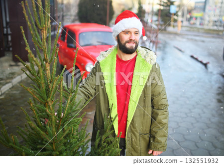 attractive bearded man in santa hat holding a christmas tree in his hands and smiling on the background of a red retro car. attractive bearded man in santa hat holding a christmas tree in his hands and smiling on the background of a red retro car. 132551932