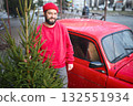 an attractive bearded man in a red sweater and hat holds a Christmas tree in his hands against the background of a red retro car. 132551934