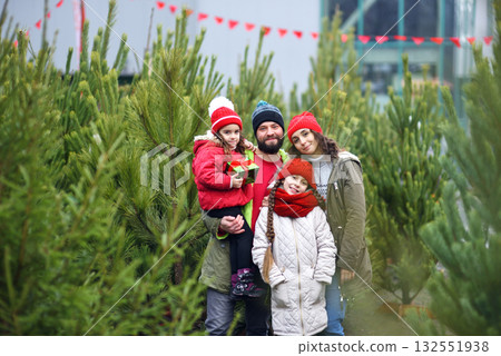 a young family - parents and two daughters are smiling against the backdrop of a Christmas tree shop. Preparing for Christmas. 132551938