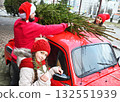 a little girl with pigtails holds a branch of a Christmas tree in her hands against the background of parents who tie a Christmas tree to the roof of a red retro car. 132551939