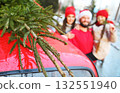 a Christmas tree on the roof of a red car against the backdrop of a happy family - mom, dad and daughter. 132551940