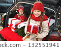 two little girls are smiling while sitting in the trunk of a car and holding Christmas gifts in their hands against the background of Christmas tree branches, a red plaid and Christmas lights. 132551941