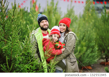 parents hold their little daughter in their arms with a Christmas present in their hands and smile against the backdrop of many Christmas trees and snow. 132551945