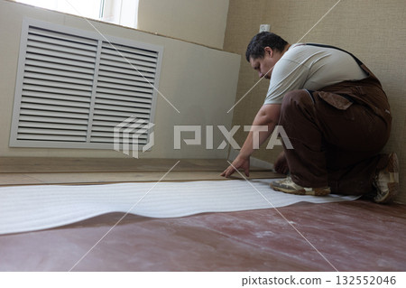 A man lays laminate flooring throughout his home. A man in work overalls lays insulation and joins laminated wood panels on a subfloor during a home improvement and renovation project. 132552046