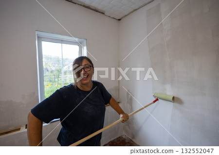 A woman smiles and laughs as she applies primer to a wall with a roller working on a home improvement and DIY project that transforms the interior space. A cheerful woman primes the walls of a house. 132552047