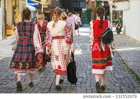 Women wearing traditional Bulgarian folk costumes walking on cobbled street during cultural event. Concept of heritage, folklore and national identity 132552079