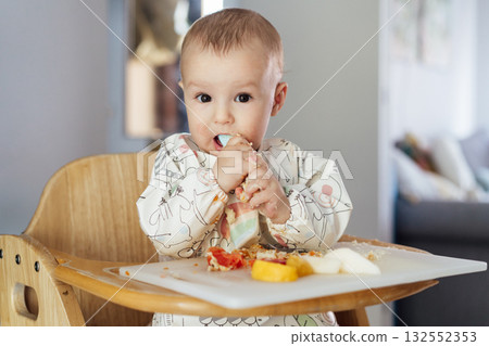 Cute baby with redness cheeks learning to eat food by himself on high chair baby led weaning or blw. Infant self-feeding solid food, introducing allergens into food. Kids' food tolerance develop. 132552353