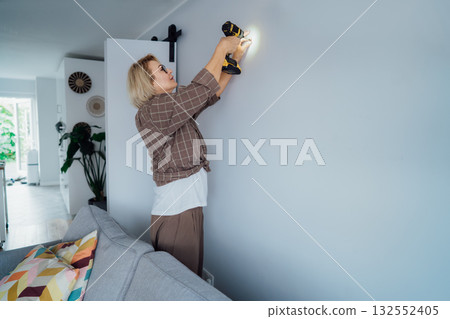 Side view middle aged woman in plaid shirt screwing a screw into the wall with electrical screwdriver. Woman wants to put a picture on the wall. Housekeeping work. Doing repair herself. DIY concept Side view middle aged woman in plaid shirt screwing a screw into the wall with electrical screwdriver. Woman wants to put a picture on the wall. Housekeeping work. Doing repair herself. DIY concept 132552405