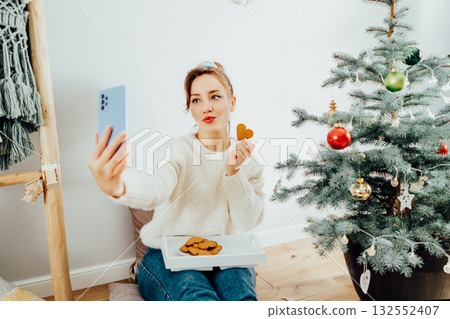 Happy Young Woman in cosy sweater taking selfie photo while relaxing on floor cusions near potted christmas tree in modern Scandi interior home. Cozy winter holidays. Content creating. Selective focus Happy Young Woman in cosy sweater taking selfie photo while relaxing on floor cusions near potted christmas tree in modern Scandi interior home. Cozy winter holidays. Content creating. Selective focus 132552407
