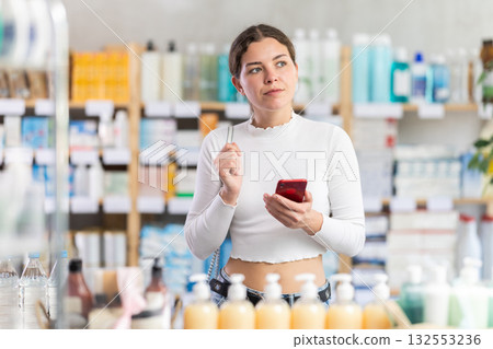 Female shopper searches for necessary goods on the shelves of a supermarket, checking the list on smartphone 132553236