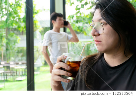 Two Thai LGBTQ friends enjoying lunch break at a cozy cafe one sipping iced coffee and the other talking on phone in a natural warm and relaxing atmosphere 132553564