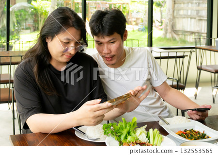 Two Thai LGBTQ friends enjoying lunch taking food photos to share on social media showing friendship happiness and real lifestyle moments in cozy daylight cafe Two Thai LGBTQ friends enjoying lunch taking food photos to share on social media showing friendship happiness and real lifestyle moments in cozy daylight cafe 132553565