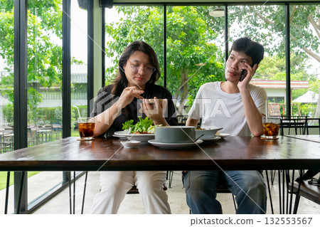 Two Thai LGBTQ friends enjoying lunch together one checking photos while the other on a phone call in a cozy cafe showing friendship relaxation and real daily life moments Two Thai LGBTQ friends enjoying lunch together one checking photos while the other on a phone call in a cozy cafe showing friendship relaxation and real daily life moments 132553567