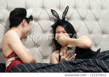 Two Thai LGBTQ friends laughing together in bed using smartphone relaxing and sharing happy evening moments showing friendship joy and authentic lifestyle before sleeping 132553615