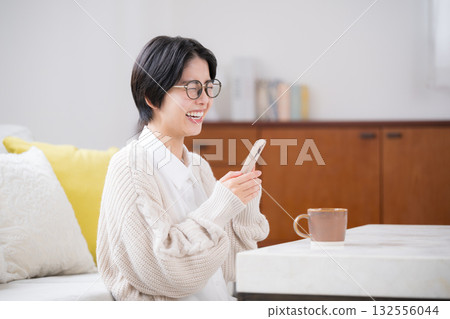 A cute smiling woman wearing a knitted sweater in her room and laughing out loud while looking at her smartphone 132556044