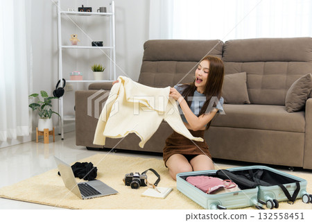 Young happy woman at home, people enjoying activity of packing for travel, sitting on rug, holding cream sweater, selecting fashion clothes, preparing suitcase, planning vacation with laptop, camera 132558451