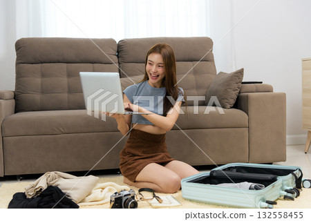 Young happy woman at home enjoying activity, planning travel, using laptop for booking vacation, packing suitcase for exciting adventure with clothes and camera 132558455