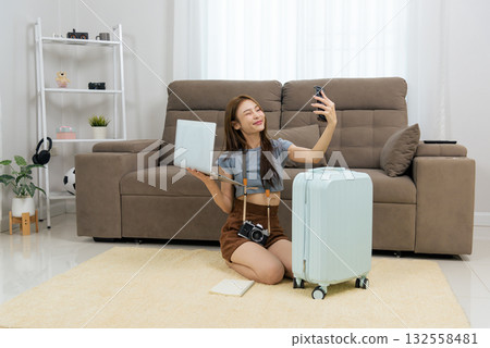 Young happy woman at home enjoying activity, planning exciting travel vacation with modern laptop and smartphone, taking selfie, packing suitcase for upcoming journey, feeling joyful leisure time 132558481
