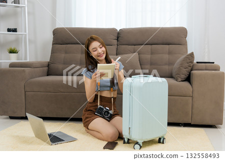 Young woman at home, happy people planning travel activity, writing ideas in notebook using pen, vintage camera, laptop, phone and suitcase nearby, excited for future journey 132558493