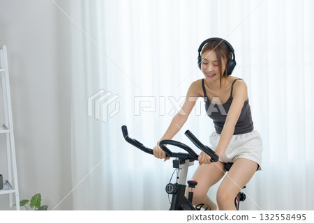Young happy woman enjoys home activity, people engage in fitness, spinning bike workout for healthy lifestyle, wearing headphones, comfortable indoor residence Young happy woman enjoys home activity, people engage in fitness, spinning bike workout for healthy lifestyle, wearing headphones, comfortable indoor residence 132558495