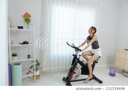 Young woman happy enjoys home activity exercising on stationary bike embracing healthy lifestyle with headphones, bright room setting displays fitness dedication for wellness goals Young woman happy enjoys home activity exercising on stationary bike embracing healthy lifestyle with headphones, bright room setting displays fitness dedication for wellness goals 132558496