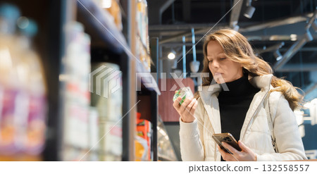 Woman studies the yogurt jar label for its ingredients and shelf life during her healthy food shopping in a grocery store aisle. Woman studies the yogurt jar label for its ingredients and shelf life during her healthy food shopping in a grocery store aisle. 132558557