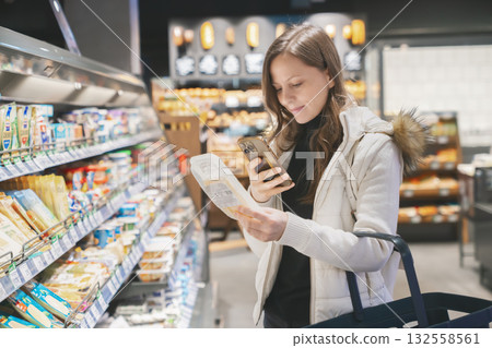 Woman examines a package of cheese in the grocery store, scanning the label with her phone to learn about its ingredients and expiration date while shopping. 132558561