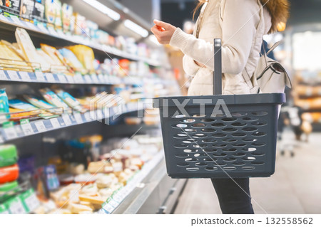 A woman stands in a grocery store aisle, holding a black shopping basket. She is carefully selecting products from the shelves filled with various food items, enjoying her shopping experience. 132558562