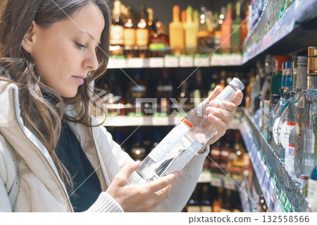 A woman with long hair examines the label on a vodka bottle while standing in the alcoholic beverages section of a supermarket. She appears focused on her choice. A woman with long hair examines the label on a vodka bottle while standing in the alcoholic beverages section of a supermarket. She appears focused on her choice. 132558566