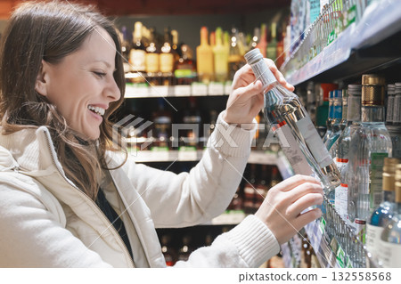 A joyful woman wearing a white jacket smiles as she reaches for a bottle of vodka in a store, preparing for a party and social event with friends. A joyful woman wearing a white jacket smiles as she reaches for a bottle of vodka in a store, preparing for a party and social event with friends. 132558568