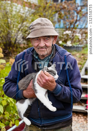 Close up portrait of elderly Russian man in autumn garden holding his gray and white cat with affection, peaceful rural lifestyle in Russia. Close up portrait of elderly Russian man in autumn garden holding his gray and white cat with affection, peaceful rural lifestyle in Russia. 132558755