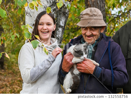 Smiling young woman and her elderly grandfather holding a gray and white cat outdoors on an autumn day, surrounded by birch trees and yellow leaves. Smiling young woman and her elderly grandfather holding a gray and white cat outdoors on an autumn day, surrounded by birch trees and yellow leaves. 132558756