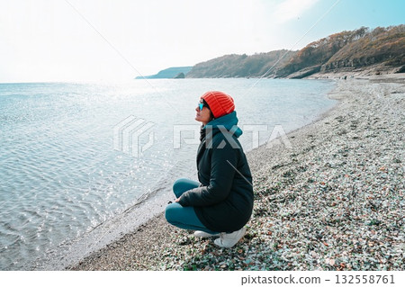 Russian woman of 50 sitting on Glass Bay beach in Vladivostok, Primorsky Krai, near the Japanese Sea, enjoying the view and harmony on a cool autumn or spring day. 132558761